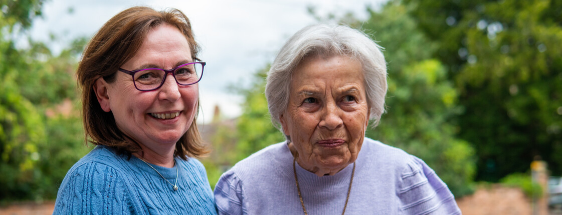 © centreforcare.ac.uk via Centre for Ageing Better Eine Frau mittleren Alters mit Brille stützt eine Senorin, die eine Halskette trägt, beim Gehen. Im Hintergrund sind grüne Bäume.