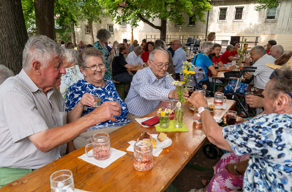 Älteres Ehepaar feiert mit Freunden im Freien in einem Garten, Getränke und Kuchen auf dem Tisch.