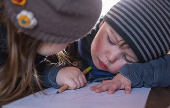 Zwei Kinder sitzen an einem Tisch, eines der beiden Kinder hat einen Stift in der Hand und sein Kopf liegt auf seiner Hand, sein Blick ist traurig zu Boden gerichtet. Beide tragen eine Mütze.