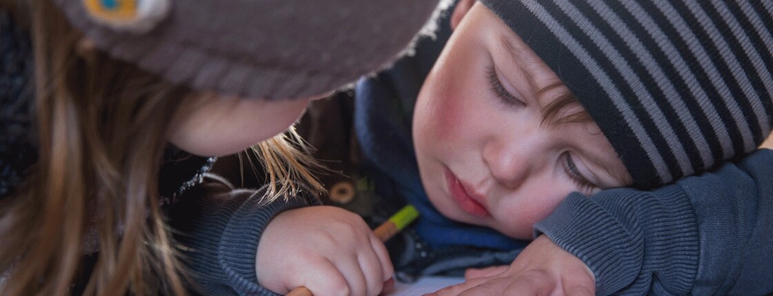 Familien in Not Zwei Kinder sitzen an einem Tisch, eines der beiden Kinder hat einen Stift in der Hand und sein Kopf liegt auf seiner Hand, sein Blick ist traurig zu Boden gerichtet. Beide tragen eine Mütze.