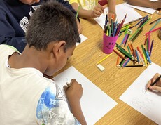 Ein Kind sitzt an einem Tisch und zeichnet mit einem Buntstift auf ein Blatt Papier, während weitere Kinder daneben ebenfalls malen; auf dem Tisch liegen viele bunte Stifte.