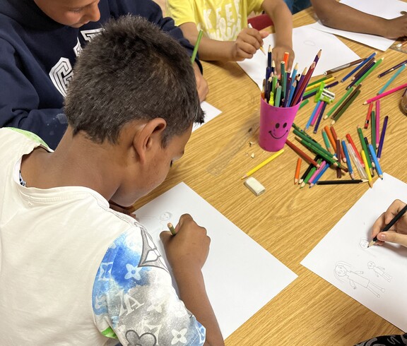 Ein Kind sitzt an einem Tisch und zeichnet mit einem Buntstift auf ein Blatt Papier, während weitere Kinder daneben ebenfalls malen; auf dem Tisch liegen viele bunte Stifte.