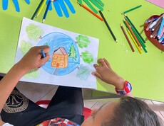 Ein Kind malt ein farbiges Bild mit einem Haus, Bäumen und blauem Himmel; auf dem Tisch liegen viele Buntstifte und bunte Papierhände.