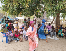 Frauen tanzen, im Kreis darum sitzen Frauen am Boden und lächeln.