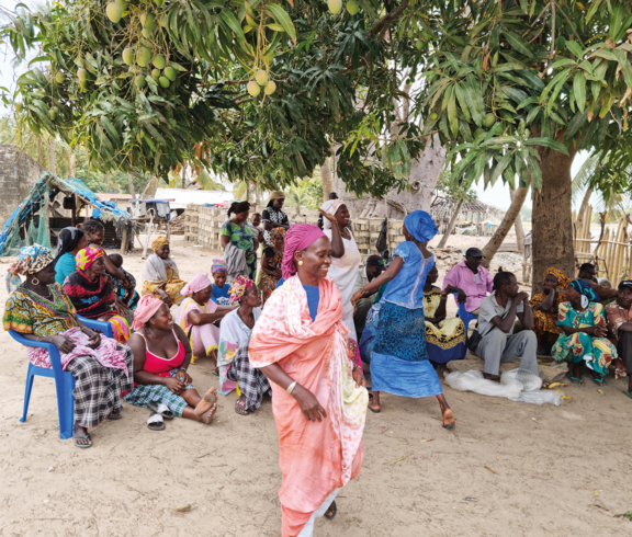 Frauen tanzen, im Kreis darum sitzen Frauen am Boden und lächeln.