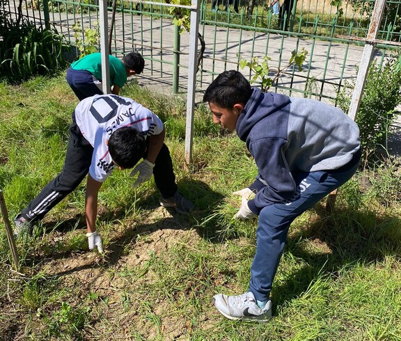 Zwei Jungen ziehen im Gras eines Gartens Unkraut, während weitere Kinder und Erwachsene im Hintergrund ebenfalls im Freien arbeiten.