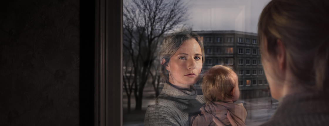 Eine Frau mit Kind am Arm schaut aus dem Fenster. Ihr Gesicht spiegelt sich im Fensterglas wider. Vor dem Fenster ist eine Wohngegend erkennbar.