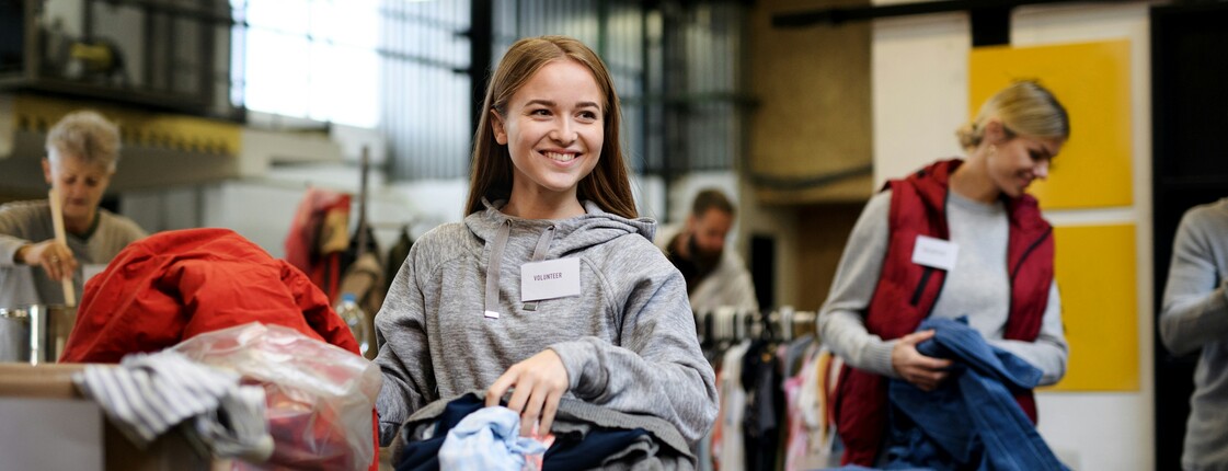 Eine lachende junge Frau sortiert Second-Hand-Kleidung in einer Sachspenden-Annahmestelle.