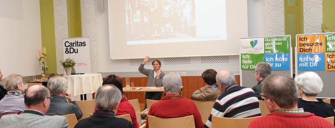 Mehrere Personen sitzen und schauen auf eine Leinwand und den Mann im Vordergrund