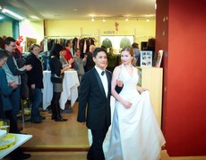 Ein junger Mann im Anzug führt mit einer jungen Frau im weißen Kleid die Kleidung der carla vor. Die Menschen hinter ihnen applaudieren.