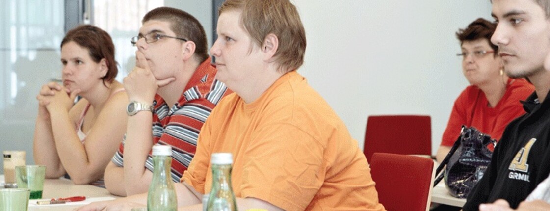 Mehrere Personen sitzen an einem Tisch, vor sich haben sie etwas zu trinken stehen und auch etwas zum schreiben.