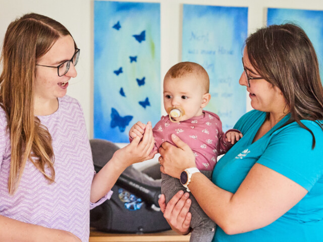 Mitarbeiterin hält Baby im Arm, Mutter steht gegenüber und gibt dem Baby die Hand..