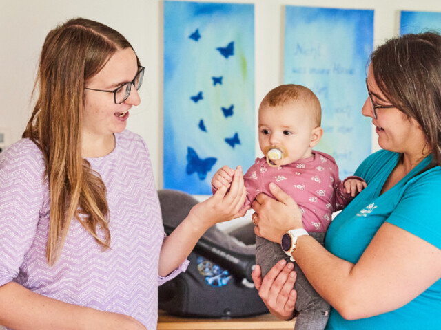 Mitarbeiterin hält Baby im Arm, Mutter steht gegenüber und gibt dem Baby die Hand..