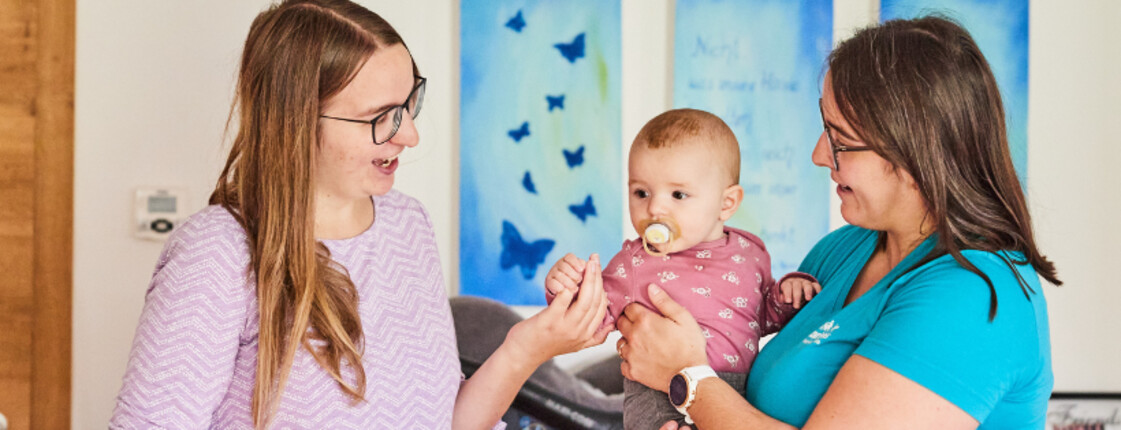 Mitarbeiterin hält Baby im Arm, Mutter steht gegenüber und gibt dem Baby die Hand..