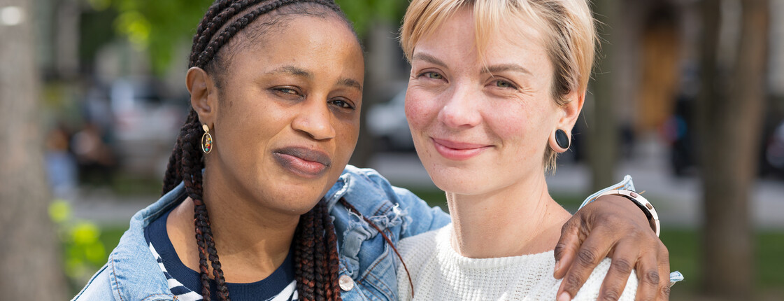 Patenschaften für Geflüchtete Zwei junge Frauen, die sich im Buddy-Projekt kennengelernt haben, stehen Arm in Arm in einem Park.