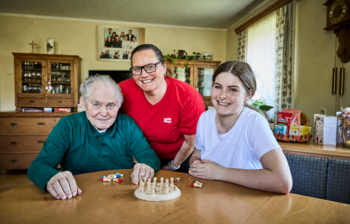 Pflegefachkraft und Schülerin sind zu Hause bei einem Kunden der mobilen Pflege. Sie sitzen mit ihm an einem Tisch uns spielen ein Brettspiel.