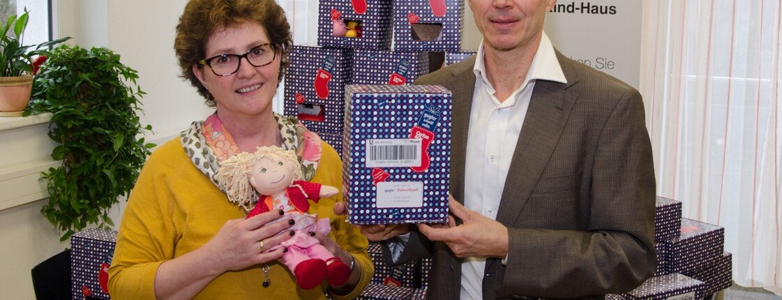 Zwei Personen halten eine Schachtel und eine Puppe in die Kamera. Hinter ihnen sind noch mehrere Schachteln.