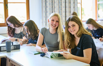 Schüler*innen sitzen im Klassenzimmer. 2 Schülerinnen lesen ein Buch über Familienarbeit und lächeln.