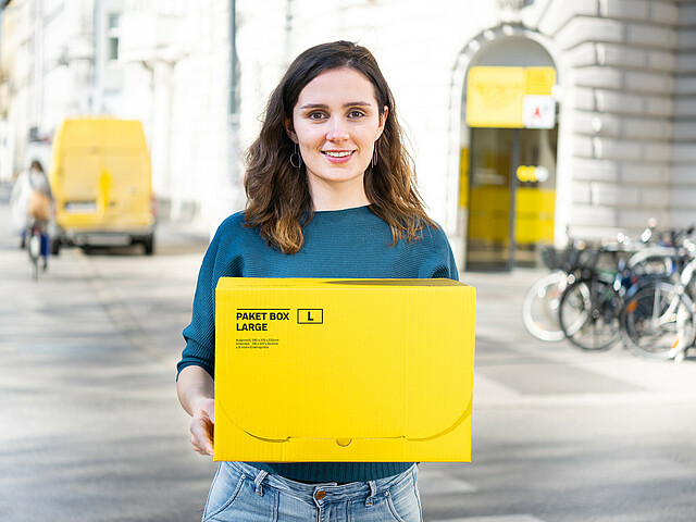 Junge Frau hält ein gelbes Postpaket, sie lächelt und schaut in die Kamera. Im Hintergrund ist eine Postfiliale.