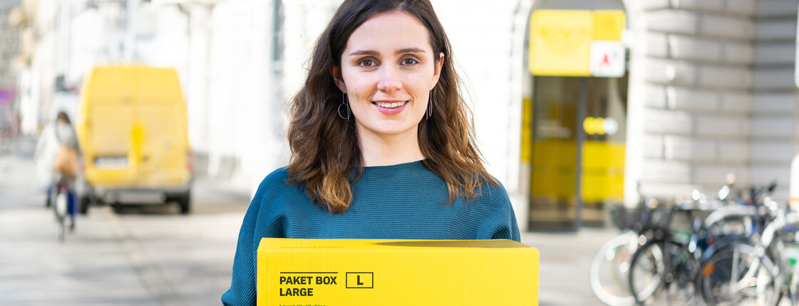 Junge Frau hält ein gelbes Postpaket, sie lächelt und schaut in die Kamera. Im Hintergrund ist eine Postfiliale.