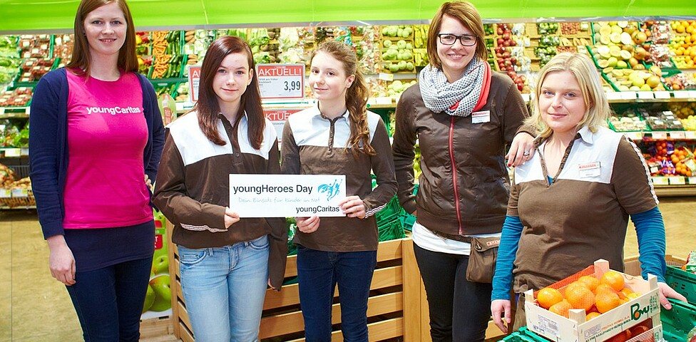 Zwei junge Mädchen stehen zusammen mit einer youngCaritas Mitarbeiterin und zwei Mitarbeiterinnen eines Supermarktes bei Gemüseabteilung.