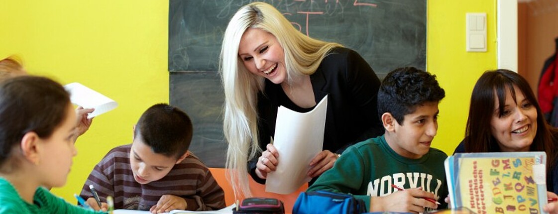 Schulkinder sitzen an einem Tisch und machen Aufgabe, daneben steht eine junge Frau mit Zetteln in der Hand.