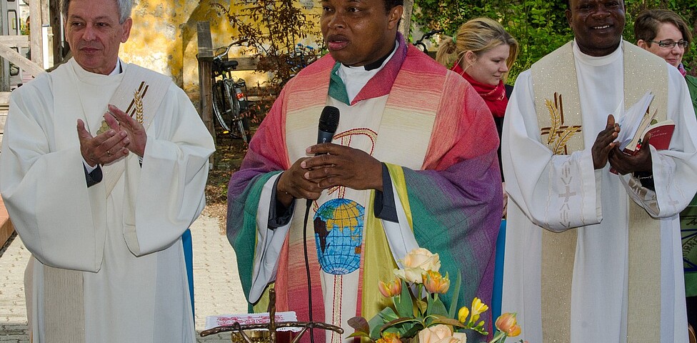 Drei Pfarrer stehen hinter einem Tisch, der mittlere trägt bunte Kleidung während die beiden anderen in Weiß gekleidet sind.