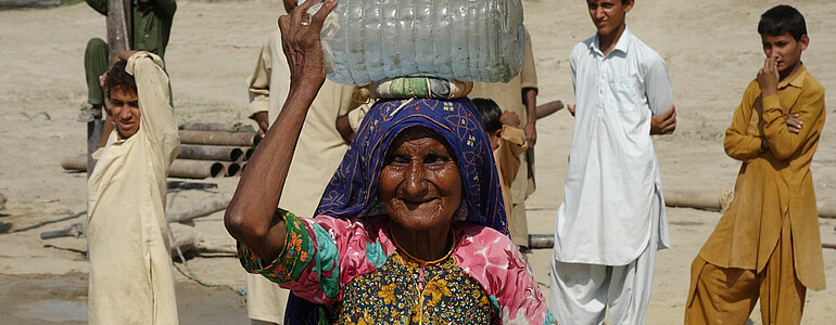 Alte Frau mit Wasserbehälter am Kopf in pakistanischer Wüste