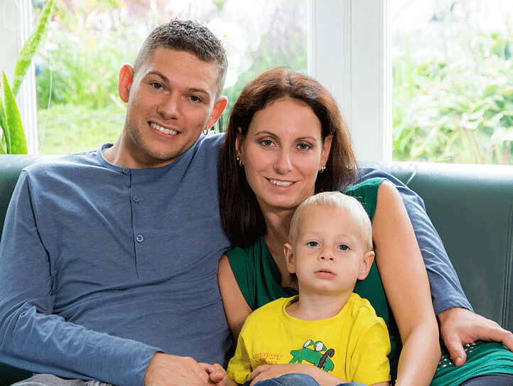 Ein kleine Familie, bestehend aus einem Vater, einer Mutter und einem kleinen Jungen, sitzen auf einem Sofa und lächeln in die Kamera.