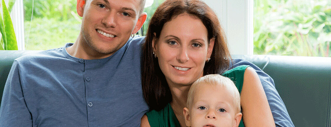 Ein kleine Familie, bestehend aus einem Vater, einer Mutter und einem kleinen Jungen, sitzen auf einem Sofa und lächeln in die Kamera.