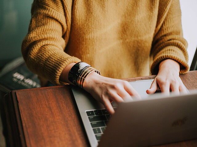 Eine junge Frau arbeitet am Laptop, ihre Hände auf der Tastatur sind im Fokus, ihr Gesicht nicht zu sehen.